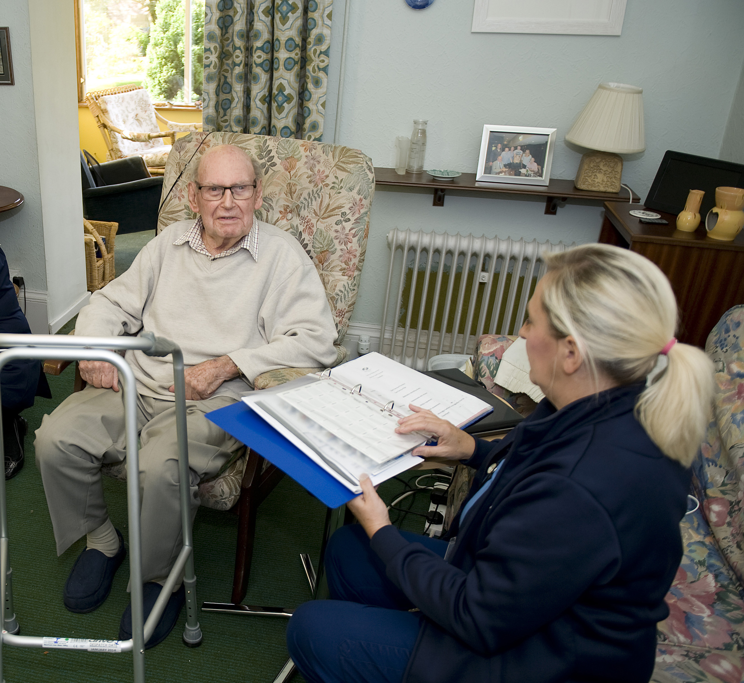  An elderly man sitting in his room with a healthcare worker looking at his notes in a folder